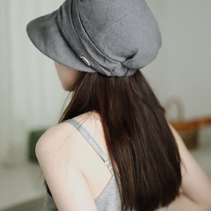 May include: A gray linen newsboy cap with a wide brim and a fold-over detail. The cap is worn by a person with long dark hair.
