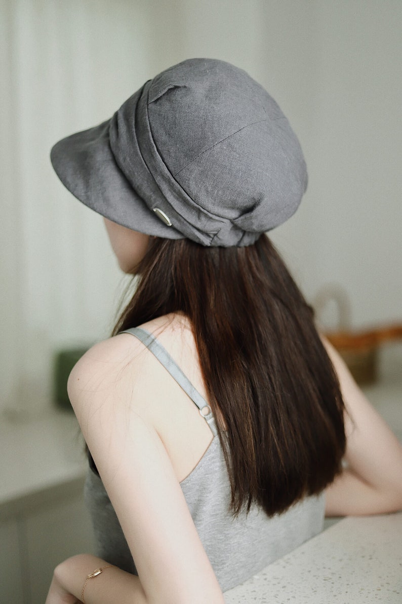 May include: A gray linen newsboy cap with a wide brim and a fold-over detail. The cap is worn by a person with long dark hair.