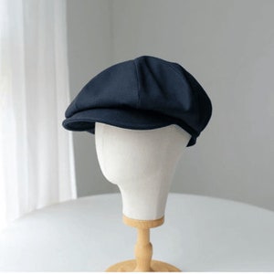 May include: A navy blue newsboy cap displayed on a white mannequin head. The cap has a rounded crown and a small, stiff brim. The background is a soft, neutral color, with a hint of a window and curtains.