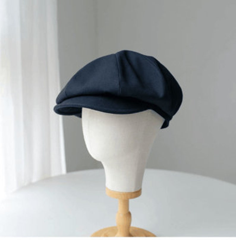 May include: A navy blue newsboy cap displayed on a white mannequin head. The cap has a rounded crown and a small, stiff brim. The background is a soft, neutral color, with a hint of a window and curtains.