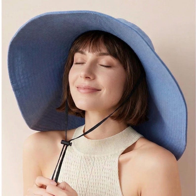 May include: A wide-brimmed, light blue sun hat with a black chin strap. The hat has a soft, slightly textured appearance. The brim is large and provides ample shade. The hat is worn with a cream-colored, sleeveless top.