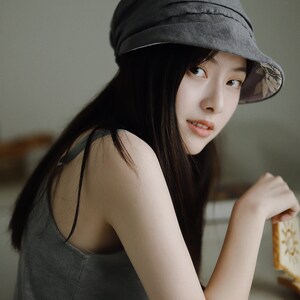 May include: A woman wearing a gray hat with a floral lining and a gray tank top. She is leaning on a counter and holding a cookie.
