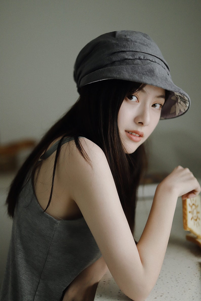 May include: A woman wearing a gray hat with a floral lining and a gray tank top. She is leaning on a counter and holding a cookie.