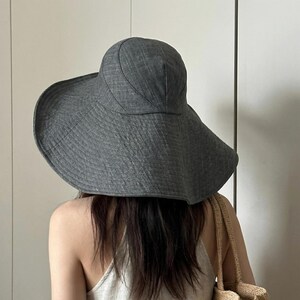 May include: A wide-brimmed, gray sun hat. The hat has a rounded crown and a broad, downward-sloping brim. The fabric appears to have a textured weave. The hat is worn by a person with long, dark hair, and a beige tote bag is visible.
