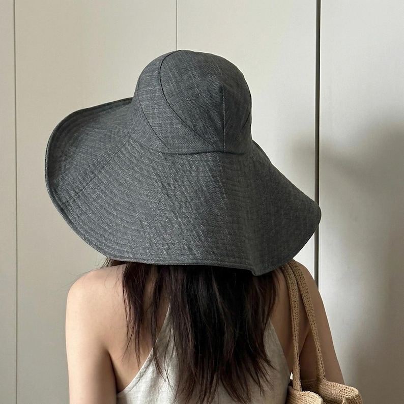 May include: A wide-brimmed, gray sun hat. The hat has a rounded crown and a broad, downward-sloping brim. The fabric appears to have a textured weave. The hat is worn by a person with long, dark hair, and a beige tote bag is visible.