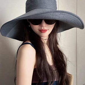 May include: A wide-brimmed, gray sun hat with a UPF50 rating. The hat features a structured crown and a broad, floppy brim. The image also shows black sunglasses.