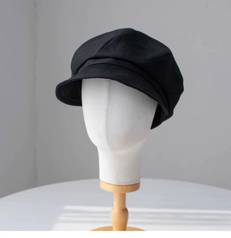 Puede incluir: Una gorra de newsboy de algod&oacute;n negro con una visera ligeramente curvada. La gorra est&aacute; en una cabeza de maniqu&iacute; blanca.