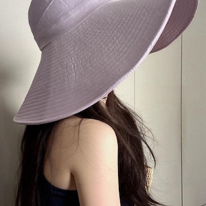 May include: A light purple sun hat with a wide brim. The hat has a textured appearance and a rounded crown. The brim is curved, providing ample shade. The hat is made of a lightweight material, suitable for sunny days.