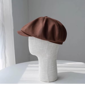 May include: A brown newsboy cap with a short brim. The cap is made of a textured fabric and is displayed on a mannequin head. The cap has a rounded crown and a small, stiff brim. The color is a rich, warm brown.