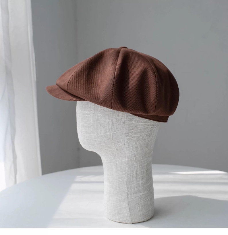 May include: A brown newsboy cap with a short brim. The cap is made of a textured fabric and is displayed on a mannequin head. The cap has a rounded crown and a small, stiff brim. The color is a rich, warm brown.