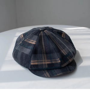 May include: A dark gray newsboy cap with a plaid pattern in shades of brown and teal. The cap has a rounded crown and a small, stiff brim. The fabric appears to be a woven material.
