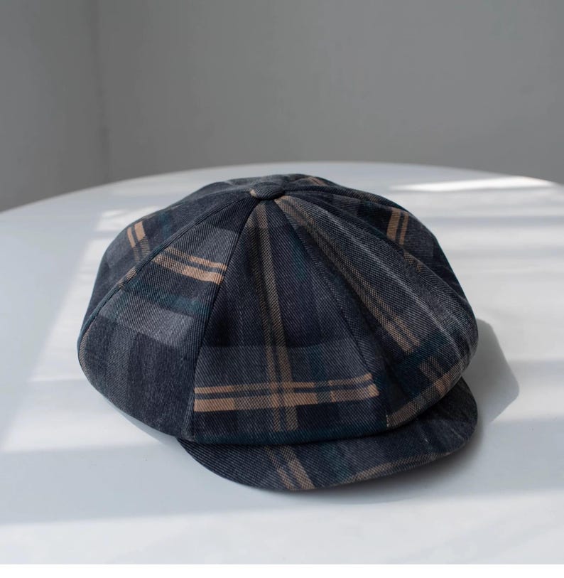 May include: A dark gray newsboy cap with a plaid pattern in shades of brown and teal. The cap has a rounded crown and a small, stiff brim. The fabric appears to be a woven material.