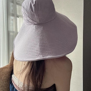 May include: A lavender sun hat with a wide brim. The hat is made of a textured fabric and has a rounded crown. The brim is designed to provide ample shade. The hat is worn by a person.