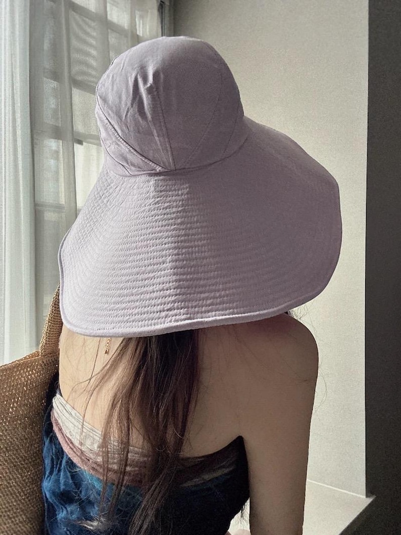 May include: A lavender sun hat with a wide brim. The hat is made of a textured fabric and has a rounded crown. The brim is designed to provide ample shade. The hat is worn by a person.