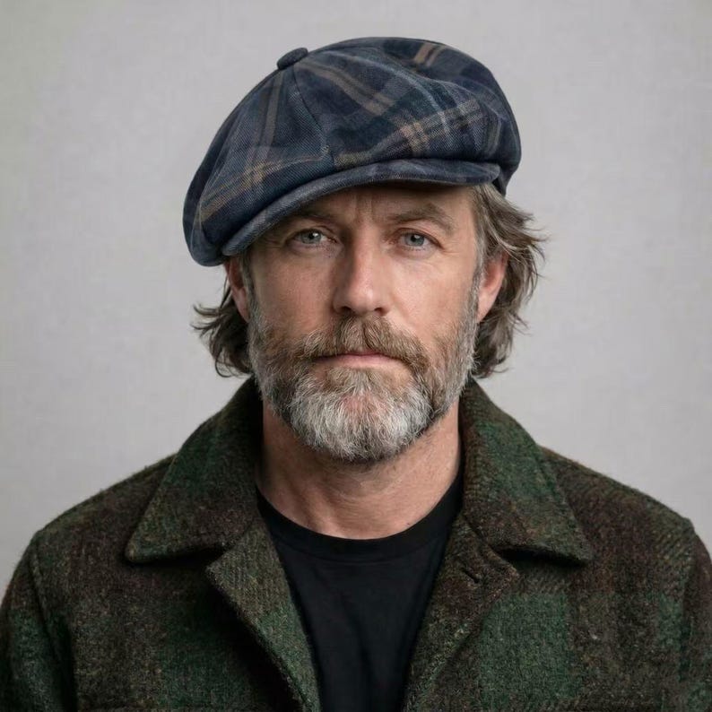 May include: A blue plaid newsboy cap with a button on top. The cap has a dark blue base with brown and tan plaid lines. The person in the image is wearing a green and brown plaid jacket and a black shirt.