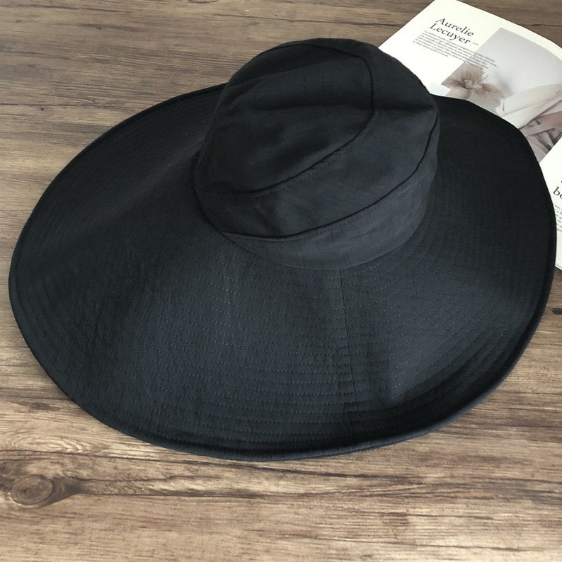 May include: A black sun hat with a wide brim and a rounded crown. The hat is made of a textured fabric and is designed to provide shade from the sun. The hat is sitting on a wooden surface.