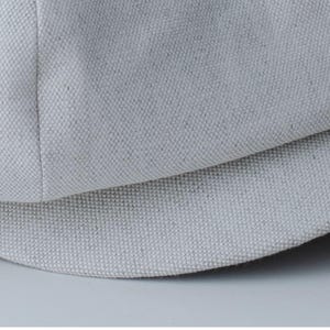 May include: Close-up of a light gray, textured fabric, possibly canvas or linen. The fabric is tightly woven with a visible grain, and the image shows a curved section, suggesting a hat or similar item.