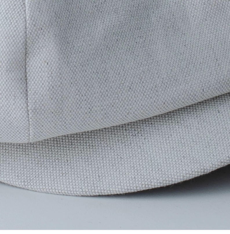 May include: Close-up of a light gray, textured fabric, possibly canvas or linen. The fabric is tightly woven with a visible grain, and the image shows a curved section, suggesting a hat or similar item.