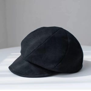 Puede incluir: Una gorra de newsboy de tela negra con una corona redonda y una visera curvada.