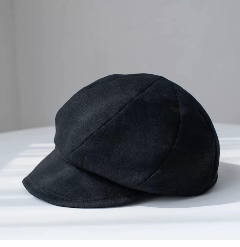 Puede incluir: Una gorra de newsboy de tela negra con una corona redonda y una visera curvada.