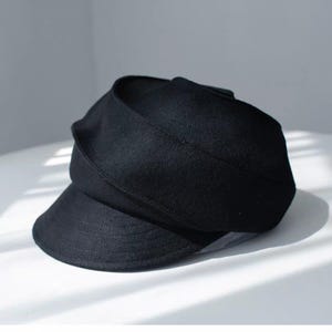 May include: A solid black cap featuring a structured crown and a wide, overlapping fabric band that wraps around the base. The hat includes a short, stiffened brim with visible horizontal stitching detail across the front surface.