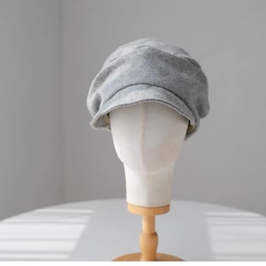 Puede incluir: Una gorra de estilo boina en color gris claro con un acabado de tela suave y texturizada. El sombrero presenta una corona redondeada de varios paneles y una visera corta y curva en la parte delantera sobre un soporte de maniquí.