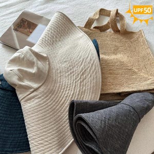 May include: A collection of sun hats and a tote bag. The hats are in beige, navy, and gray. The beige hat has a wide brim and a smaller, rounded crown. The tote bag is woven in a natural tan color. The image includes a magazine with the word "inside".