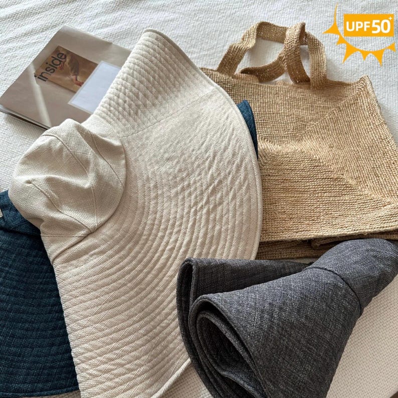 May include: A collection of sun hats and a tote bag. The hats are in beige, navy, and gray. The beige hat has a wide brim and a smaller, rounded crown. The tote bag is woven in a natural tan color. The image includes a magazine with the word "inside".