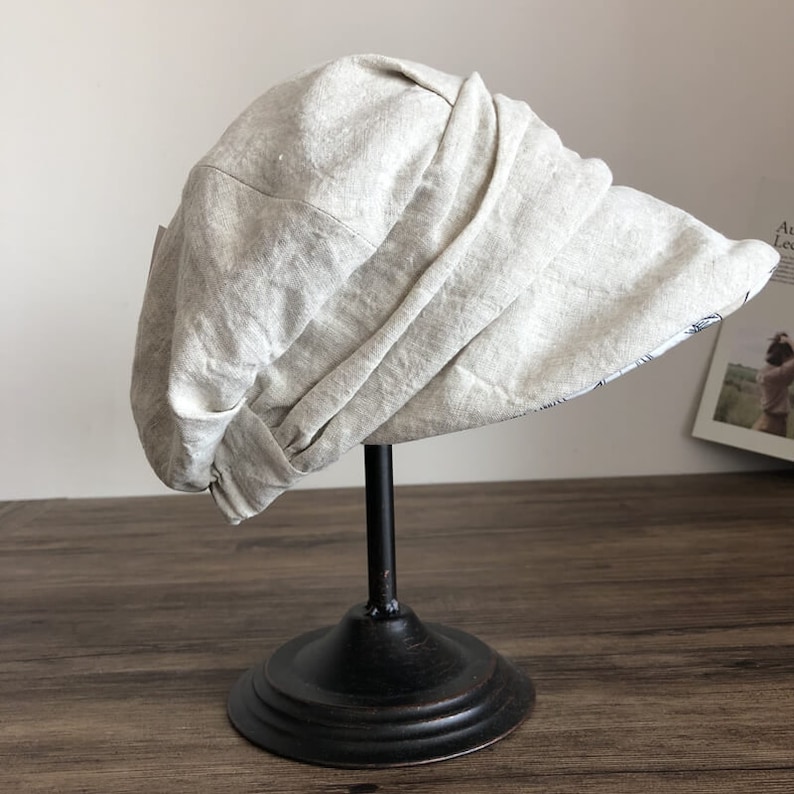 May include: A light beige linen sun hat with a wide brim, displayed on a black stand.