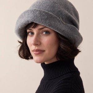 May include: A grey felted cloche hat with a rounded crown and a wide, turned-up brim. The hat features a small, delicate silver-toned metal chain detail attached to the side of the brim, adding a subtle metallic accent to the textured grey fabric.