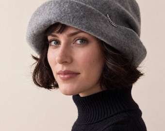 Puede incluir: Un sombrero tipo cloche de fieltro gris con una copa redondeada y un ala ancha vuelta hacia arriba. El sombrero presenta un pequeño y delicado detalle de cadena de metal plateado sujeto al lateral del ala, añadiendo un sutil toque metálico.