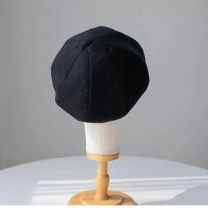 Puede incluir: Un gorro de boina negro en un soporte de madera. El gorro est&aacute; hecho de una tela suave y texturizada.