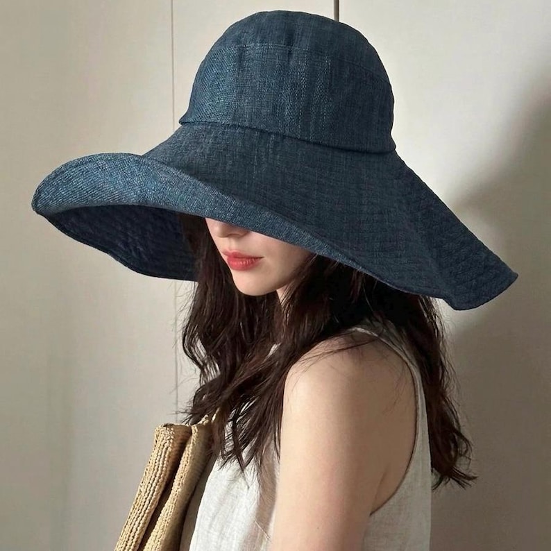 May include: A wide-brimmed, denim-blue sun hat. The hat has a rounded crown and a wide, floppy brim. The brim is slightly curved, providing ample shade. The hat is made of a textured fabric, and the color is a deep, saturated blue.