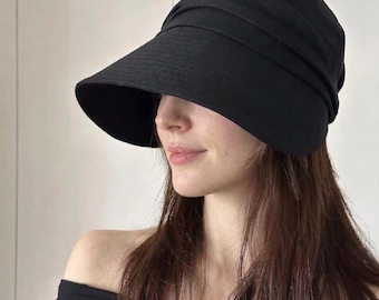 May include: A black hat with a wide brim and a gathered crown. The hat is made of a soft, textured fabric and is worn at an angle. The hat is a stylish accessory.