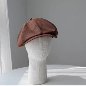 May include: A brown newsboy cap with a rounded crown and a small brim. The cap is displayed on a mannequin head. The fabric has a subtle ribbed texture. The hat is a solid, warm brown color.