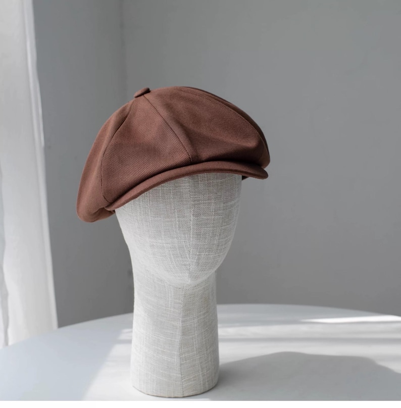 May include: A brown newsboy cap with a rounded crown and a small brim. The cap is displayed on a mannequin head. The fabric has a subtle ribbed texture. The hat is a solid, warm brown color.