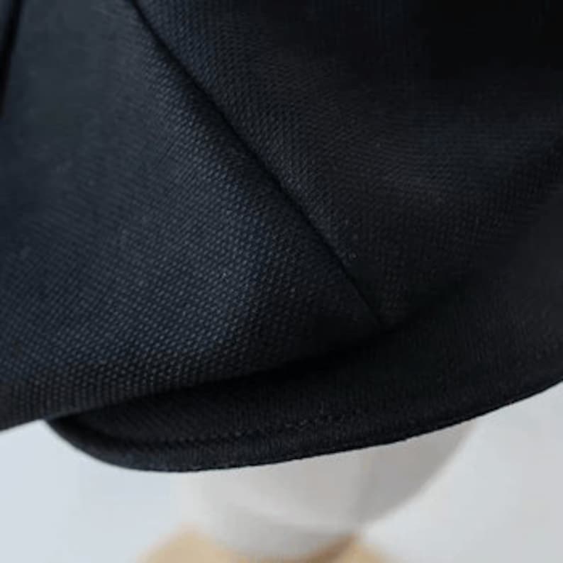 May include: Close-up of a black hat with a wide brim. The hat is made of a textured fabric and has a subtle sheen. The brim is slightly curved, and the hat appears to be well-constructed.