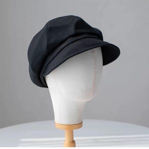 Puede incluir: Una gorra de newsboy de tela negra con una visera ligeramente curvada. La gorra est&aacute; en una cabeza de maniqu&iacute; blanca.