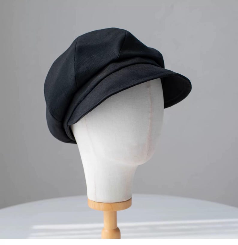 Puede incluir: Una gorra de newsboy de tela negra con una visera ligeramente curvada. La gorra est&aacute; en una cabeza de maniqu&iacute; blanca.