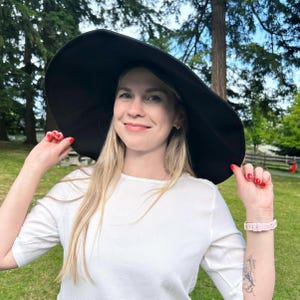 May include: A large, black sun hat with a wide brim. The hat is worn by a person wearing a white shirt. The background features green grass and trees, suggesting an outdoor setting. The person is smiling and holding the hat.