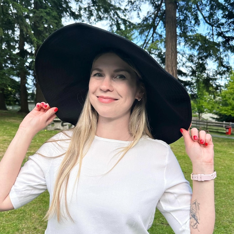 May include: A large, black sun hat with a wide brim. The hat is worn by a person wearing a white shirt. The background features green grass and trees, suggesting an outdoor setting. The person is smiling and holding the hat.