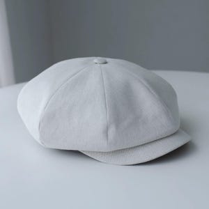May include: A light gray newsboy cap with a rounded crown and a small, stiff brim. The cap is made of a textured fabric and features a button detail at the top. This classic style is suitable for various outfits.