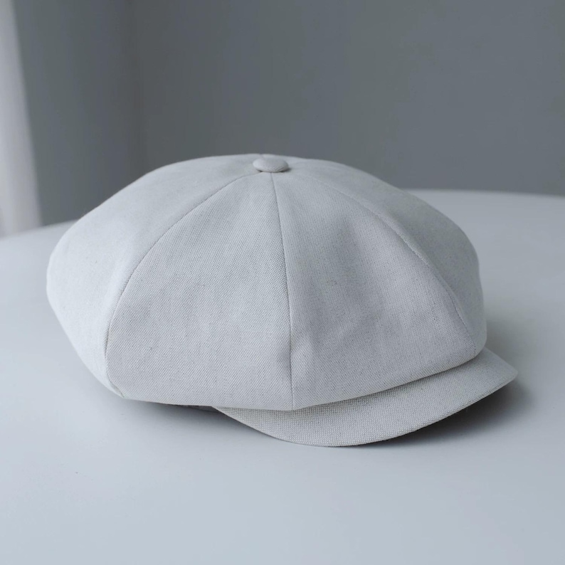 May include: A light gray newsboy cap with a rounded crown and a small, stiff brim. The cap is made of a textured fabric and features a button detail at the top. This classic style is suitable for various outfits.