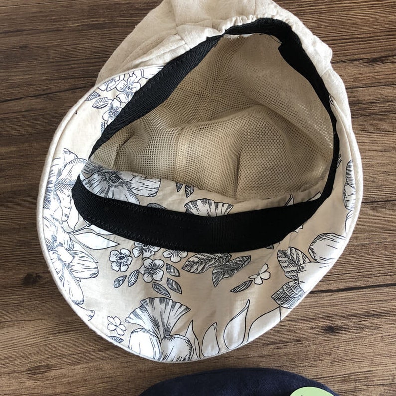 May include: A beige linen cap with a black elastic band and a floral print lining. The cap has a mesh lining.