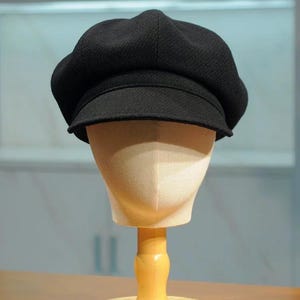 Può includere: Un berretto nero in stile newsboy con una corona arrotondata e una visiera corta e rigida. Il berretto è esposto su una testa di manichino con un supporto in legno. Il cappello ha un aspetto strutturato ed è realizzato in tessuto scuro.