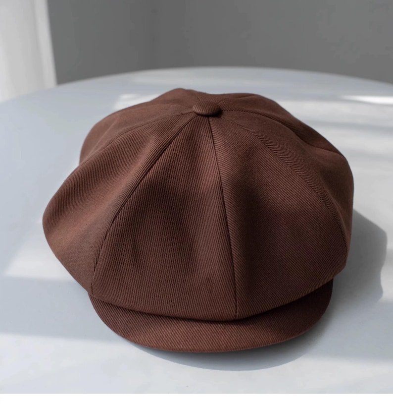 May include: A brown newsboy cap with a rounded crown and a small, stiff brim. The cap is made of a textured fabric and features a button detail at the top. This classic style is suitable for various outfits.