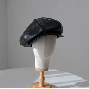 Op de afbeelding: Zwarte leren newsboy cap met een afgeronde kroon en een kleine, stijve klep. De pet wordt getoond op een witte mannequinhoofd met een houten standaard, tegen een neutrale achtergrond. De pet heeft een knoopdetail.