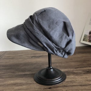 May include: A gray linen newsboy cap with a wide brim and a metal accent on the side. The cap is displayed on a black stand.