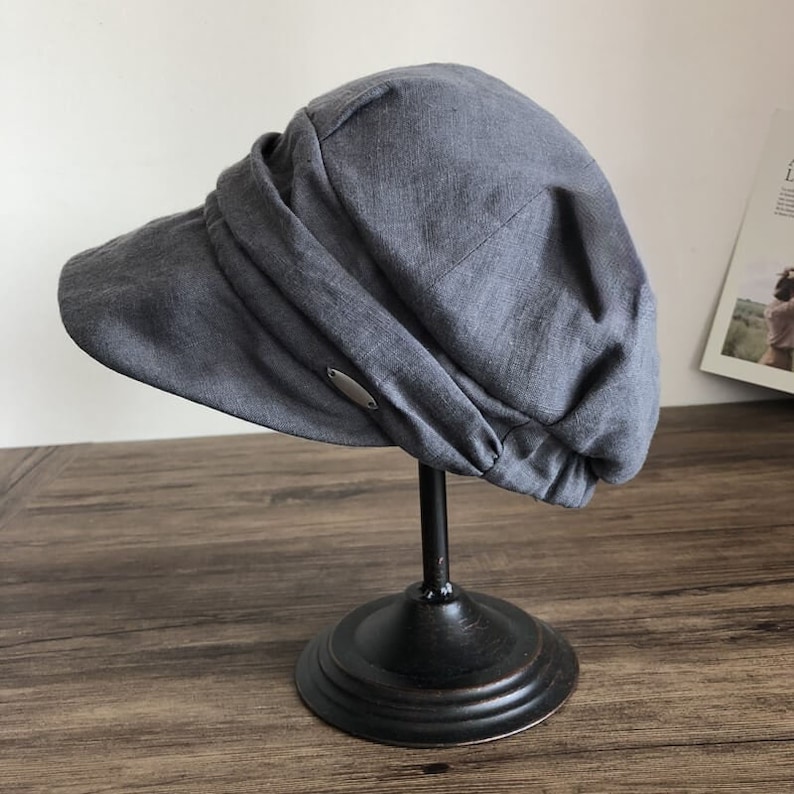May include: A gray linen newsboy cap with a wide brim and a metal accent on the side. The cap is displayed on a black stand.