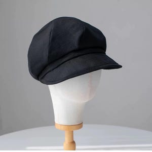 Puede incluir: Una gorra de newsboy de tela negra con una visera ligeramente curvada. La gorra est&aacute; en una cabeza de maniqu&iacute; blanca.